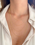 A close-up of a person's neck and upper chest wearing five layered delicate 14k yellow gold Figaro chain necklaces over a white collared shirt.