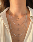 Close-up of a person's neck and upper chest wearing five layered 14k white solid gold herringbone chain necklaces of different lengths, labeled 16", 18", 20", 22", and 24".