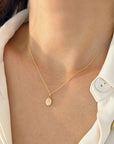 A close-up of a model wearing a delicate gold rope chain necklace with a small oval La Milagrosa Miraculous Medal pendant, styled with a white collared shirt.