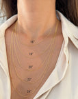 Close-up of a person's neck and upper chest wearing five layered 14k gold figaro necklaces in different lengths, labeled 16", 18", 20", 22", and 24".