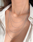 A close-up of a model's neck and collarbone wearing a layered white gold figaro necklace with five delicate chains draped in a graduated pattern over a white satin shirt.