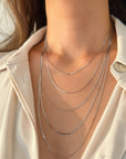 A close-up of a person wearing a layered necklace set with five delicate white gold flat snake chains draped over a cream-colored collared shirt.