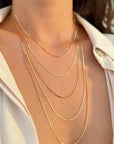 A close-up of a person's neck and upper chest wearing five layered 14k yellow solid gold herringbone necklaces of varying chain lengths, styled with a white collared shirt.