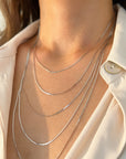 A close-up of a person's neck and upper chest wearing a layered set of five delicate white gold snake necklaces over a light cream-colored collared shirt.