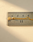 A gold rope chain necklace with a small round Guardian Angel charm pendant resting on a metal ruler to show scale, photographed on a light beige background.